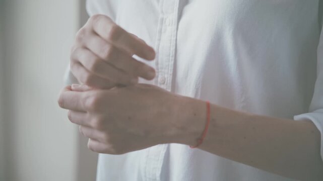 Close up of adult hands tying red string bracelet on left wrist indoors, long sleeve white shirt, minimal jewelry accessory detail, soft natural light