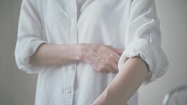 Close up of person rolling up sleeves in white button down shirt indoors, red string wrist bracelet visible, minimalist style with soft neutral light