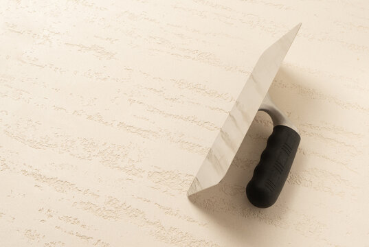 A stainless steel plastering trowel with black rubber handle resting on a travertine effect decorative plaster surface with diagonal vein relief pattern in warm beige tones.