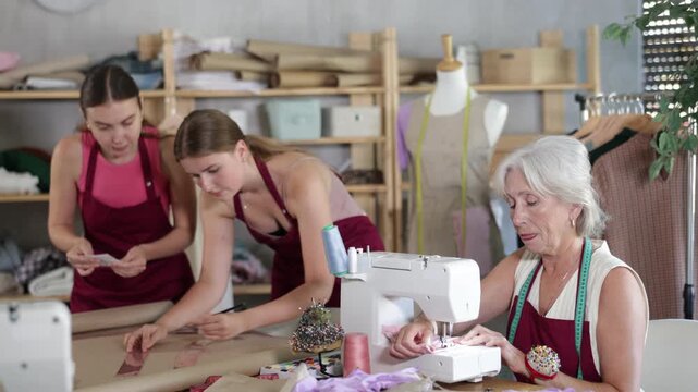 Family business, production of clothes- tailor showroom. Mature head of cloth manufacturing sewing, two female assistant drawing paper dress pattern by curves