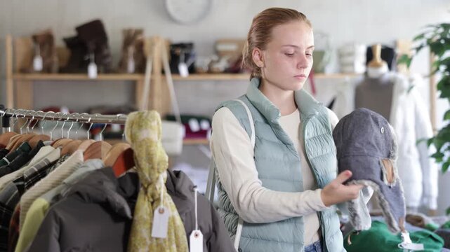  While shopping, teen girl browsing goods items and choose elect fur hat in store, buyer consider some items. Concept of seasonal wardrobe shopping. Client and holiday shopping.