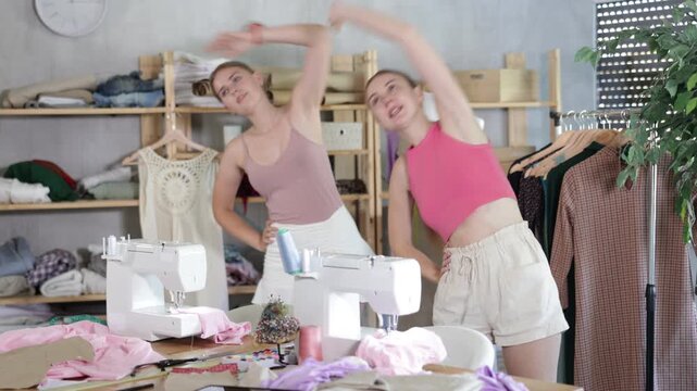 Two female dressmakers doing a warm-up for the muscles. Seamstresses rest and warm up during a break, they stand in the workshop against the background of racks with fabrics