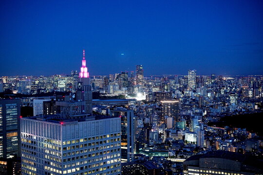 夕暮れの東京の街並みを高層ビル群が彩るパノラマ夜景。ピンク色に輝くNTTドコモ代々木ビルが都会の夜空にそびえ立つ。