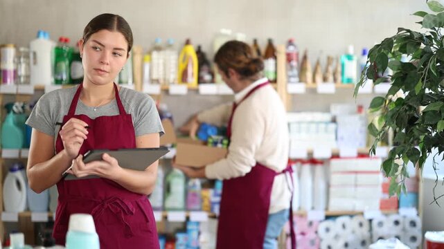 Girl seller, with tablet in hands, make inventory in store, man employee in background. Seller counts number of liquid powder bottles, records data in report, write down availability of goods in stock