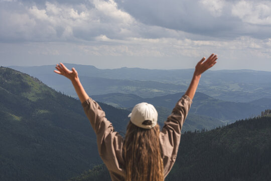 Young woman raising arms in celebration facing panoramic forested mountain landscape under cloudy sky, outdoor freedom and hiking achievement concept.