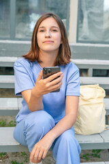 Fototapeta premium young girl wearing blue casual clothing sitting on stairs holding smartphone looking away smiling with cream colored backpack beside her outdoors during daytime candid student lifestyle portrait