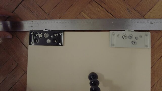 Topdown closeup installing wardrobe door roller hands using screwdriver and metal ruler to align and secure bottom roller bracket on light wood cabinet panel over parquet floor,