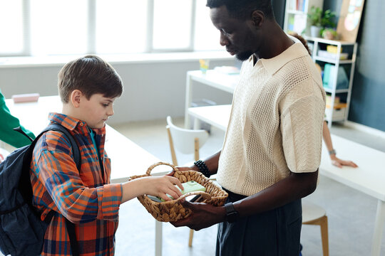 Black male teacher holding basket while schoolboy placing smartphone inside during classroom check in, supporting device rules and student focus. Useful for education policy campaigns
