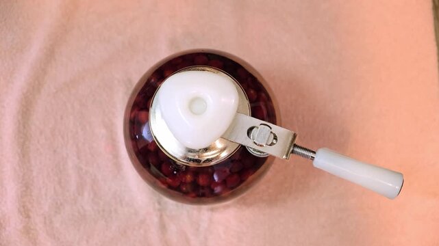 A Can Seamer. Canning cherry compote using a manual seaming machine to seal a large glass jar with metal lid. Home food preservation, traditional kitchen, winter storage preparations. 4K footage.