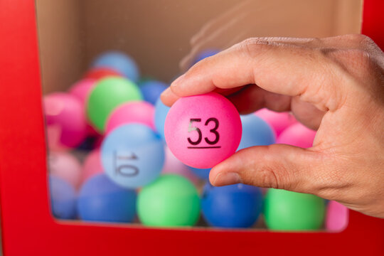 Gros plan sur une main tenant une boule de tirage au sort pour le loto, le num&eacute;ro 53. En arri&egrave;re-plan, une boite transparente avec des autres boules.