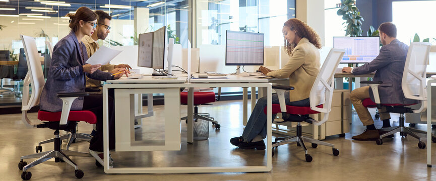 Finance team analysts and accountants in office. Computers display charts and data as reports are prepared at shared desks in a bright workspace. Finance collaboration and analytics concept.