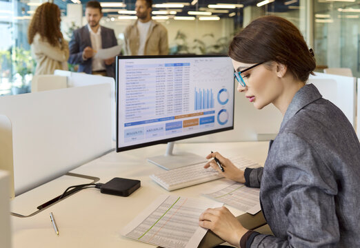 Businesswoman accountant reviews financial investment spreadsheet data. As an analyst she works on a computer in a bright office, checking charts and reports. Finance analysis and planning theme.