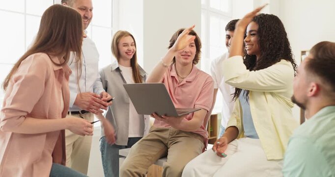 Students and teacher mark project success with celebration. A cheerful group around a laptop in classroom shares a highfive to show teamwork. Concept of achievement and collaborative learning.