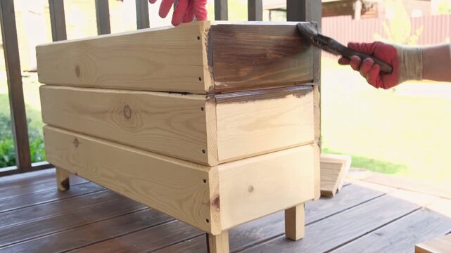 Brown oil for protection of wood by staining. Painting fence for protecting and pests. Woodworker applying finish to pine board box. Repairman varnishing with brush wooden fencing wall plank surface.