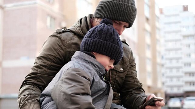 Caring father showing smartphone screen to little son outdoors. Dad and boy sitting near house, using mobile phone together. Digital Parenting, Screen Time Management. Parent and child with smartphone