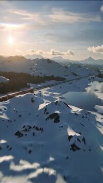 Vertical video: Descending drone tracking twin tracks over snowy coast, sun revealing red train