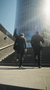 Vertical video of Caucasian and African American young adult businessmen walking up stairs toward modern office building. Footage is suitable for corporate recruitment and career growth promos