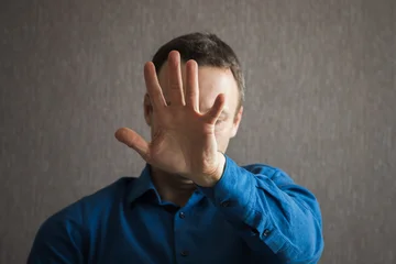 Man in blue shirt raises his hand toward the camera in a clear stop gesture, conveying privacy, refusal, avoidance and emotional distance in a portrait-style indoor setting © evannovostro
