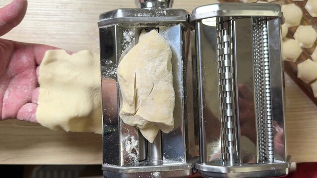 Hands feed soft dough into a metal pasta roller on a wooden counter, close view of home cooking prep, warm practical kitchen mood