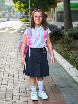Portrait of a happy little schoolgirl in uniform with glasses, ready for school. Cheerful student standing outdoors on her way to class. Back to school and education concept.