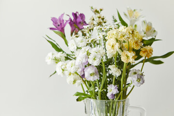 Obraz premium Fresh mixed bouquet of purple alstroemeria and white yellow stock flowers in a clear glass jug. Spring floral arrangement on a light background.