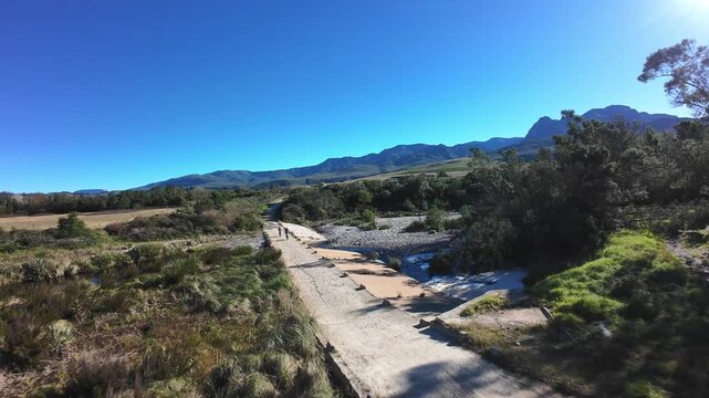 Aerial drone shot of two cyclists mountain biking over a bridge crossing a lush river in a beautiful valley
