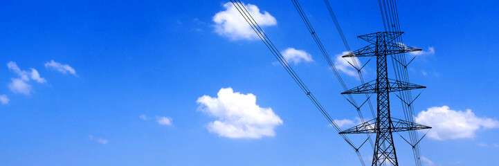 A power line tower stands tall against a bright blue sky with scattered white clouds. The silhouette of the tower and the cables create a striking contrast with the expansive sky ©  2026stockx