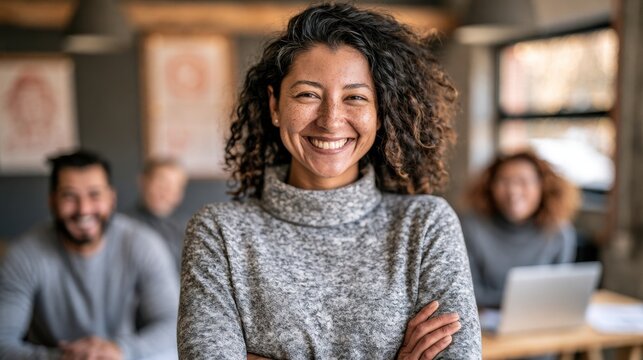 Confident young woman with curly hair smiling while standing in modern office space with crossed arms and diverse coworkers working in background