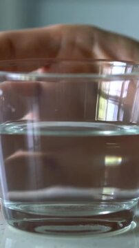 Vertical video: Lifting hand tilting half-full water glass on table showing window glow and specks