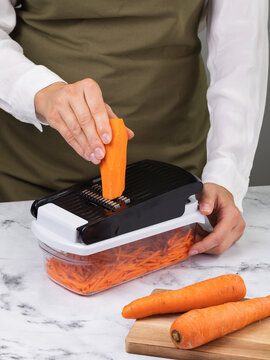 Woman in apron shredding fresh carrot into julienne strips on handheld vegetable slicer grater with container on marble countertop in kitchen healthy food preparation