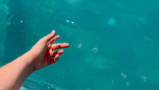 Outstretched hand reaches toward clear turquoise water, gentle ripples and light reflections create a calm summer mood, minimal composition suggests travel and relaxation