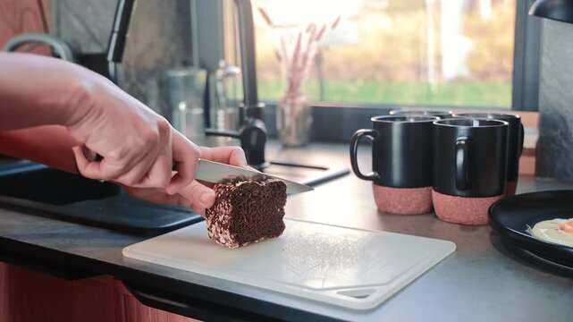 Hands slicing dark whole grain bread beside fried eggs and black mugs, Close kitchen video shows breakfast preparation in warm natural morning light