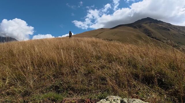 4K A hiker in a wide-brimmed hat stands in a golden grass field beneath a mountain peak with clouds drifting across a blue sky, Georgia.