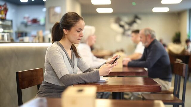 Young woman holding a mobile phone in hands while sitting at a table with a cup of coffee in a cozy cafe. Remote worker communicates with customers on a mobile phone . High quality 4k footage