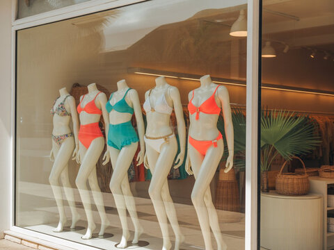 A boutique window display, a large glass case with several mannequins in bright summer bikinis, on the mannequins are women's swimsuits in coral, turquoise, and cream colors
