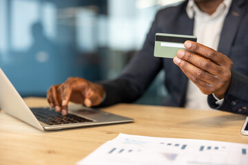 Fototapeta premium Businessman's hands holding a credit card and typing on a laptop keyboard, representing secure online banking, e-commerce, and digital transaction concepts with financial documents on the desk