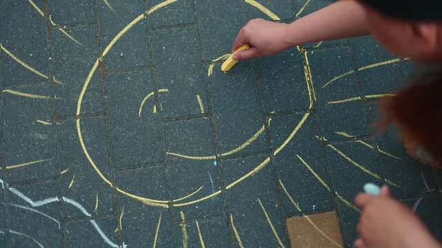 Woman draws a smiling sun on the dark pavement with yellow chalk. Childish street art creation on the sidewalk. Creative outdoor activity during a sunny day.