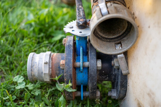 Industrial butterfly valve with manual lever handle and metal pipe flanges connected to a liquid tank on green grass.