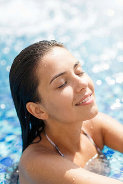 Happy beautiful young brunette woman with wet hair relaxing in a refreshing outdoor swimming pool. Smiling girl enjoying warm summer sun with closed eyes and bright blue sparkling water bokeh.