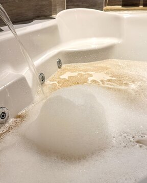 White bathtub filled with foamy bubbles and warm water, indoor bathroom setting with tiled wall, calm spa like mood for relaxation and self care