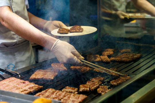 busy chef grilling meat patties on a metal grill in a restaurant kitchen close-up shot of the cooking process with plastic tongs and paper plate visible objects.
