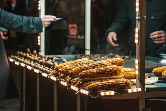 grilled corn being served at a food stall at night with warm and inviting atmosphere close-up from the customer's perspective, yellow and brown colors, metal and plastic materials, street.