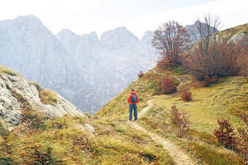 Fototapeta premium Solo hiker entering high alpine terrain beneath limestone walls along the Volusnica–Talijanka trail in Prokletije, Montenegro. Illustrates route progression and elevation gain during autumn hiking.