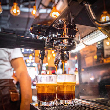 Aromatic espresso flows into two glass cups from a professional espresso machine in a warm, cozy cafe, evoking the rich morning energy and artisanal quality of gourmet coffee.