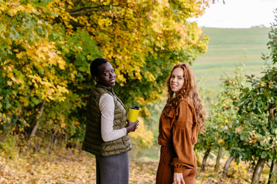 Smiling multiracial female friends standing together in autumn orchard during seasonal walk