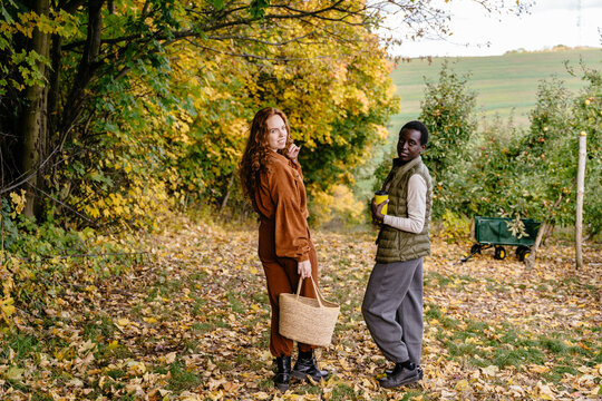 Cheerful multiracial female friends walking through autumn orchard with basket and travel mug