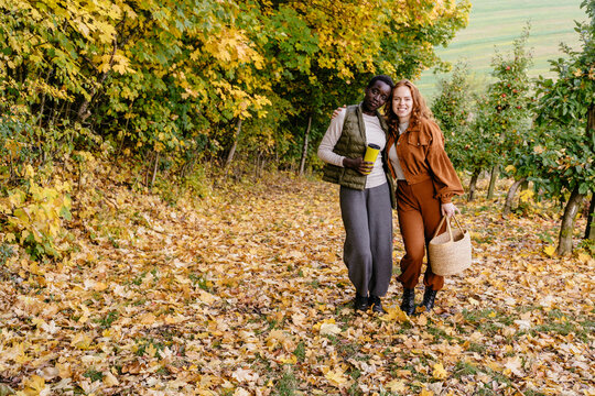 Happy multiracial female friends embracing in golden autumn orchard with basket and travel cup