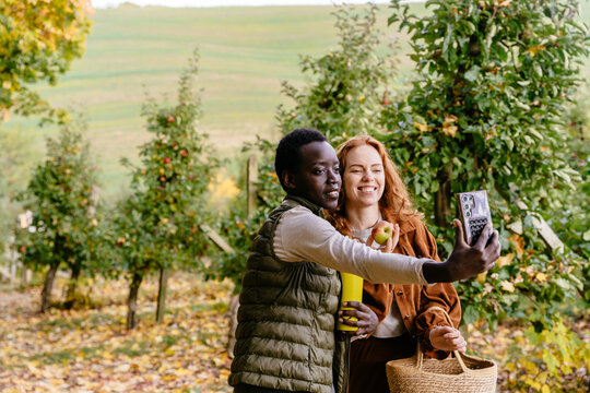 Happy multiracial female friends taking smartphone selfie with fresh apple in autumn orchard