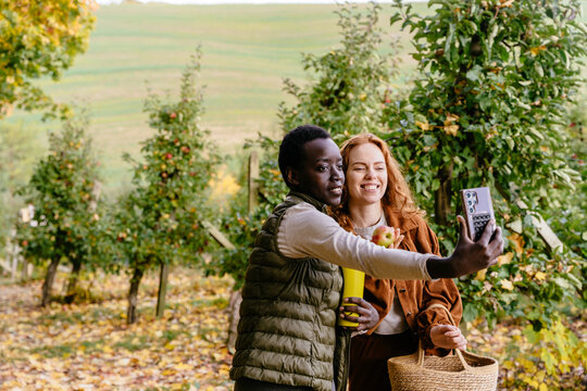 Cheerful diverse women making smartphone selfie during apple picking trip in autumn orchard