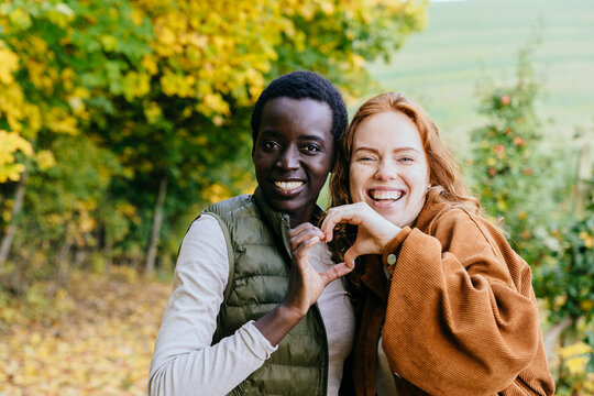 Joyful multiracial female friends making heart gesture together in colorful autumn orchard